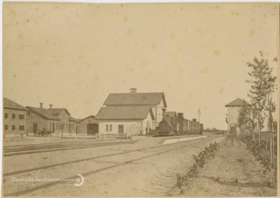 Et iltog mod København holder på stationen, set mod sydøst. Overfor stationen ligger kroen med sin rejsestald (opført 1877) og tv. et privat pakhus. Bag træet th. ses stationens vandtårn.